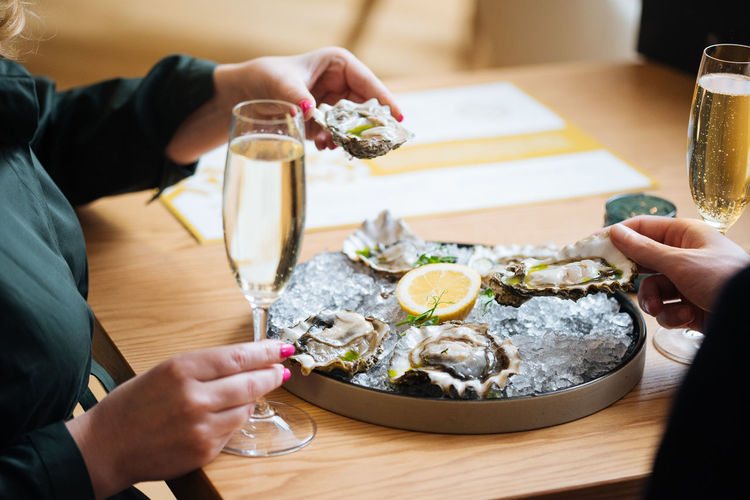 Unrecognizable couple with glasses of champagne trying delicious oysters with lemon and herbs in restaurant