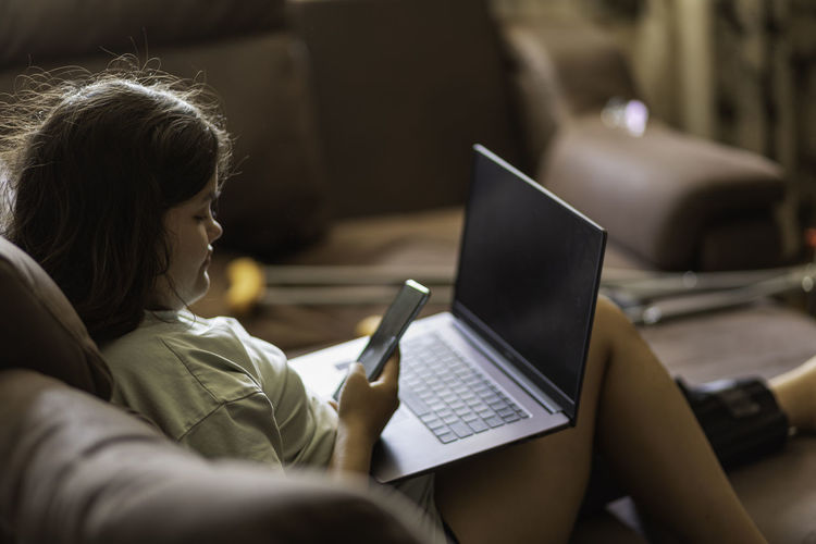 Chubby asian teen girs with injured leg and crutches talking on the phone and laptop at home