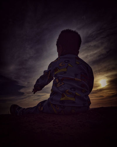 Rear view of boy sitting on land against sky during sunset