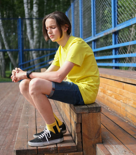 Teenager boy sitting on stairs on  playground with eyes closed.  yellow t-shirt. long haircut,