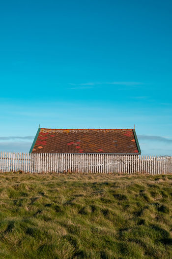 The roof of a building on the coastline appears above an old white fence against a clear blue sky. 