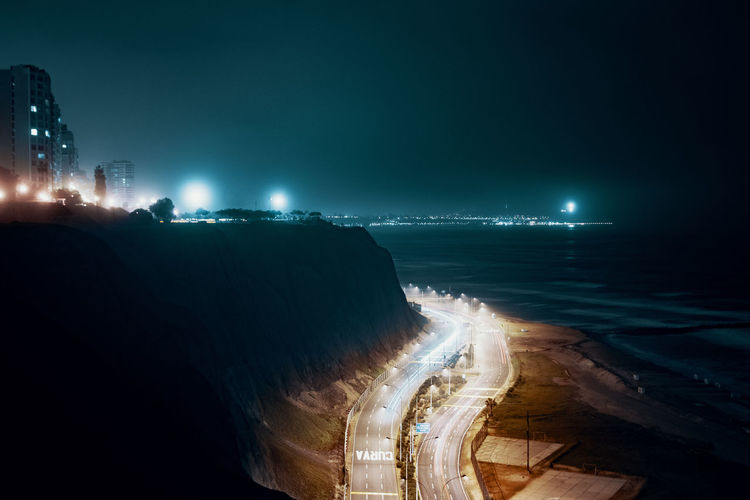 Illuminated city street by sea against sky at night