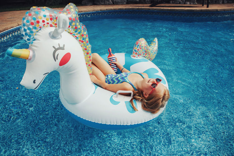 Cute adorable girl in sunglasses with drink lying on inflatable ring unicorn. kid child enjoying 