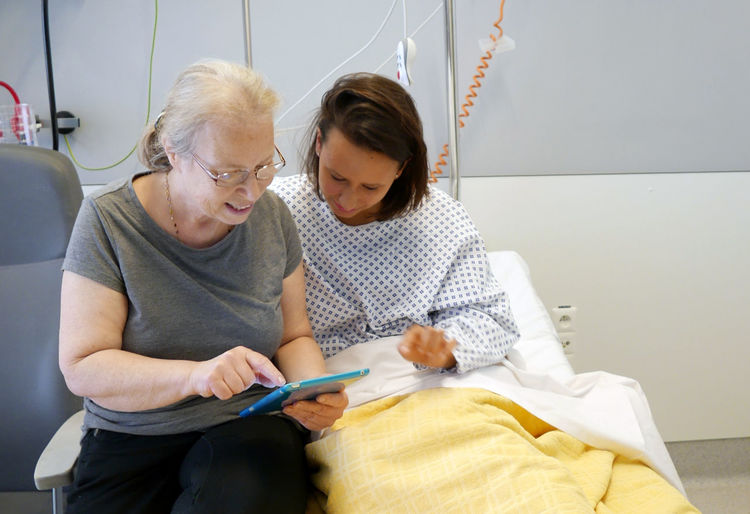 Mature woman showing digital tablet to patient while sitting on bed at hospital