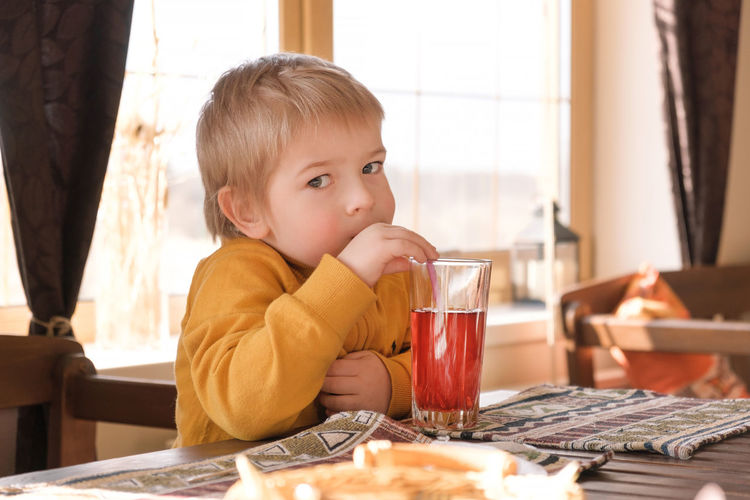 Child boy drinking lemonade in cafe. 3 years old kid drinks red juice from glasses through straws