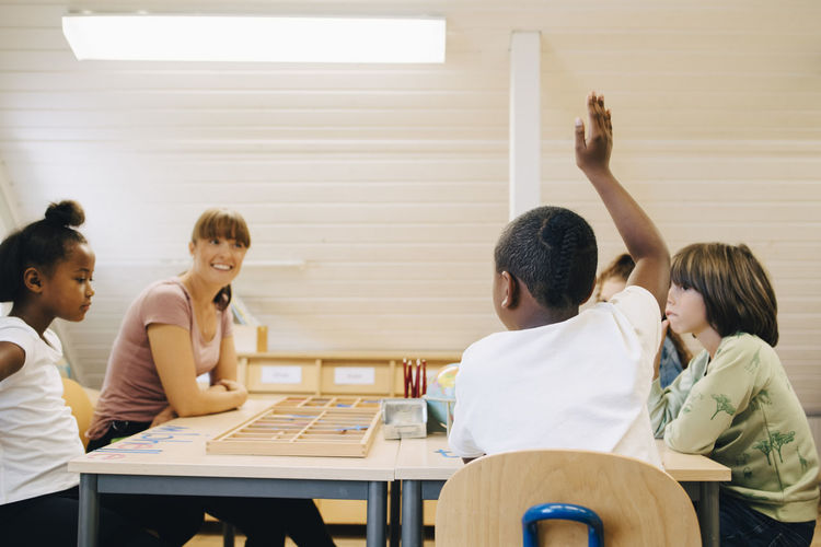 Teacher looking at boy raising hand while answering in classroom