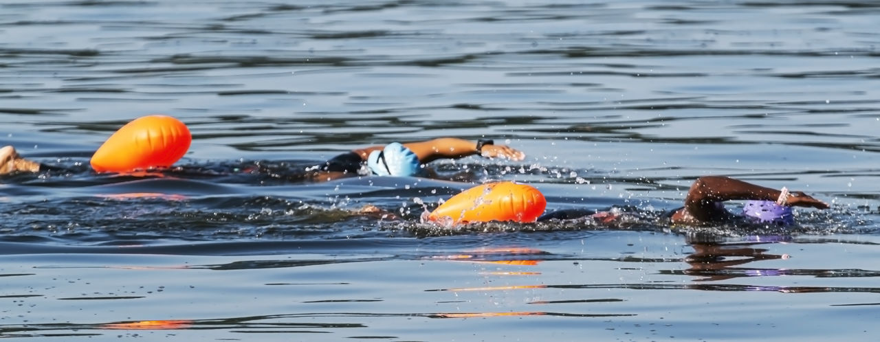 Two black triathletes are swimming together with orange safety buoys.