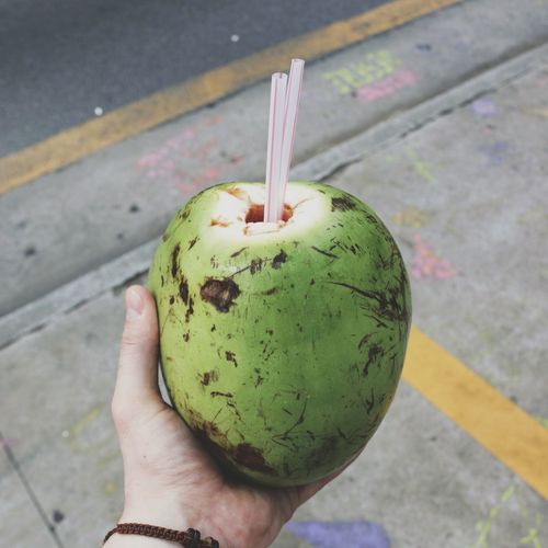 Cropped image of hand holding coconut with straws on sidewalk