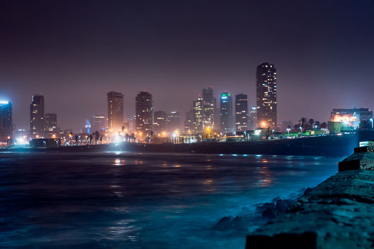 Illuminated buildings by sea against sky at night