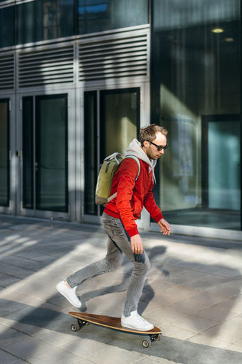 Stylish hipster in red jacket, sunglasses, sneakers and backpack riding on longboard.