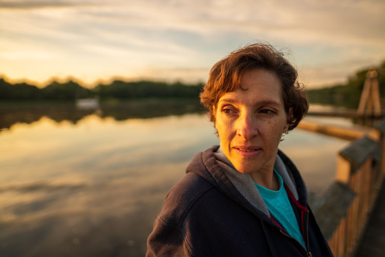 Thoughtful woman standing by lake against sky during sunset