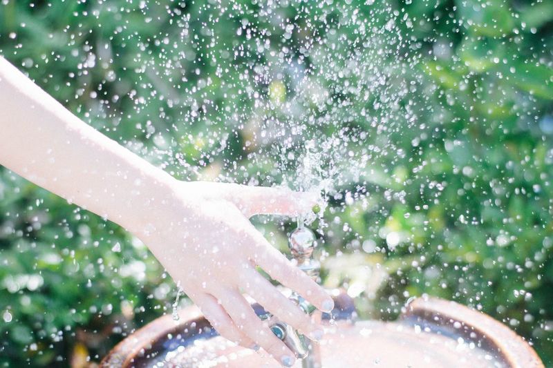 Cropped hand holding faucet spraying water during sunny day