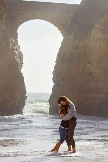 Mother and daughter embracing while standing by rock formation at beach
