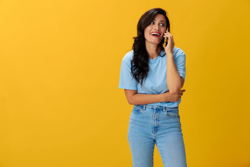 Young woman using mobile phone while standing against yellow background