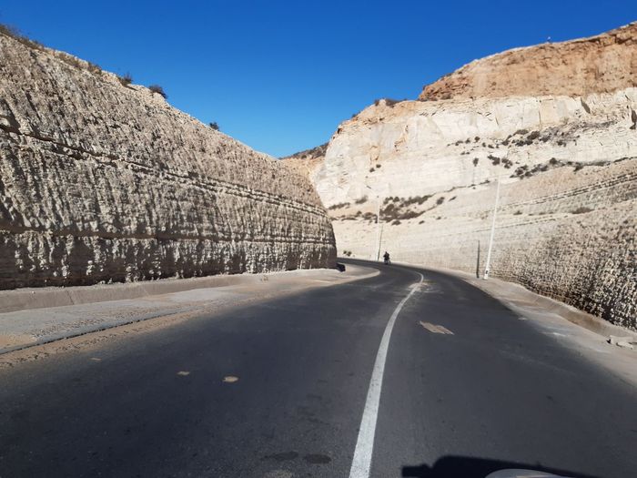 Empty road along rocky mountains against blue sky