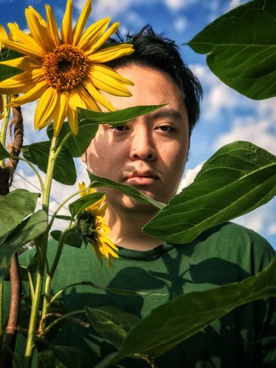Close-up portrait of a young man behind sunflower against cloudy sky.