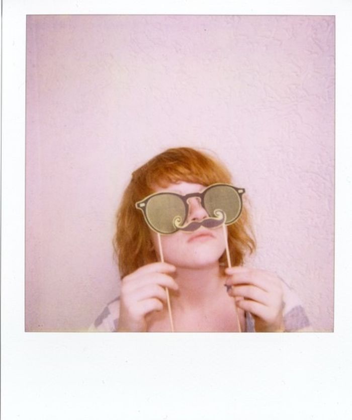 Young woman holding paper glasses and mustache against wall