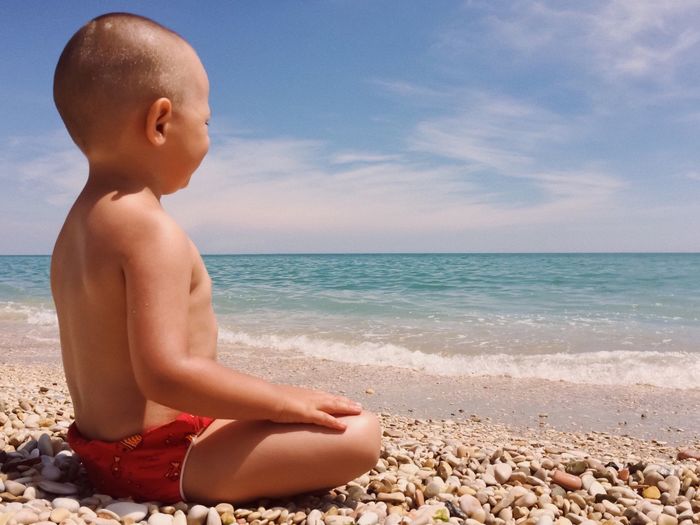 Shirtless child sitting on shore at beach during sunny day