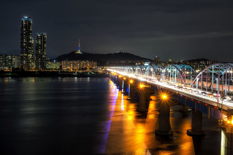 Illuminated bridge over river against buildings at night