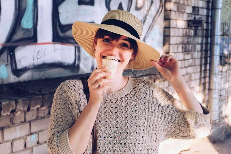 Portrait of woman in hat eating ice cream against wall