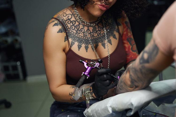 Tattoo master in gloves using professional tattoo machine while painting tattoo on arm of woman in modern tattoo studio