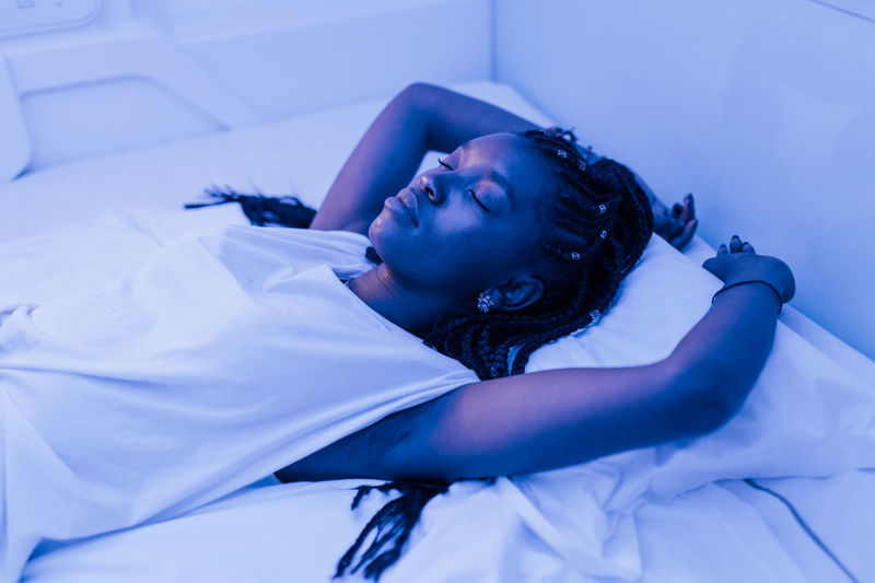 Young black woman sleeping on bed in contemporary capsule hotel with futuristic interior design and blue illumination