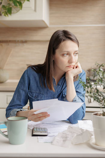 Sad young woman with documents sitting at table at home