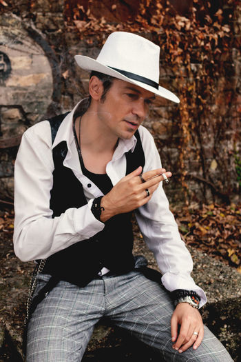 Pensive vintage styled man with fedora hat smoking a cigarette outdoors.