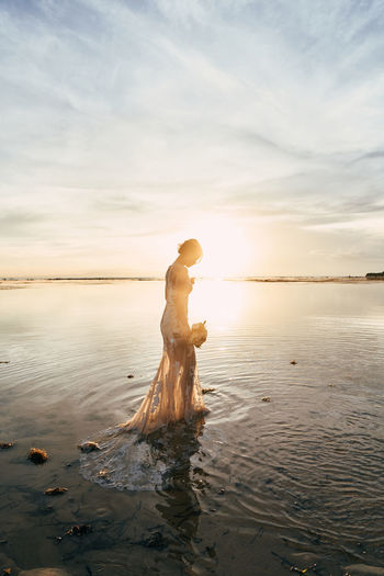 Side view of bride standing at beach against sky during sunset
