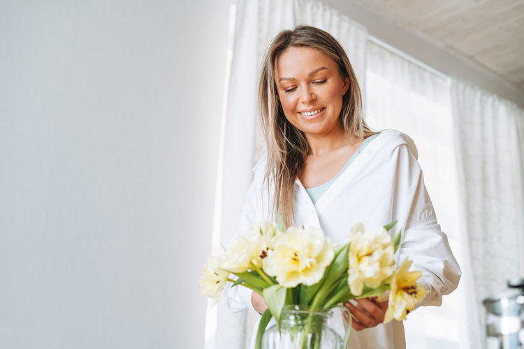 Young smiling woman with blonde long hair in white shirt with bouquet of yellow flowers at home