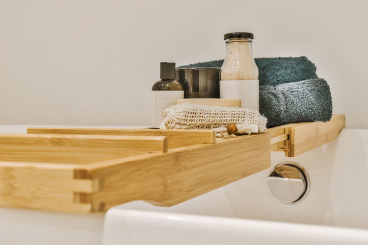 Towel and bottles with essential skincare products placed on wooden tray on white ceramic bathtub in light bathroom in apartment