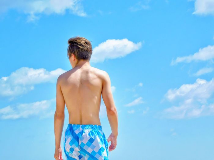 Rear view of shirtless teenage boy standing against blue sky