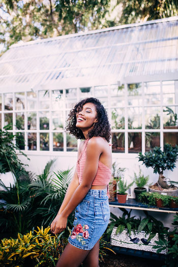 Side view of smiling young woman standing against plants at greenhouse