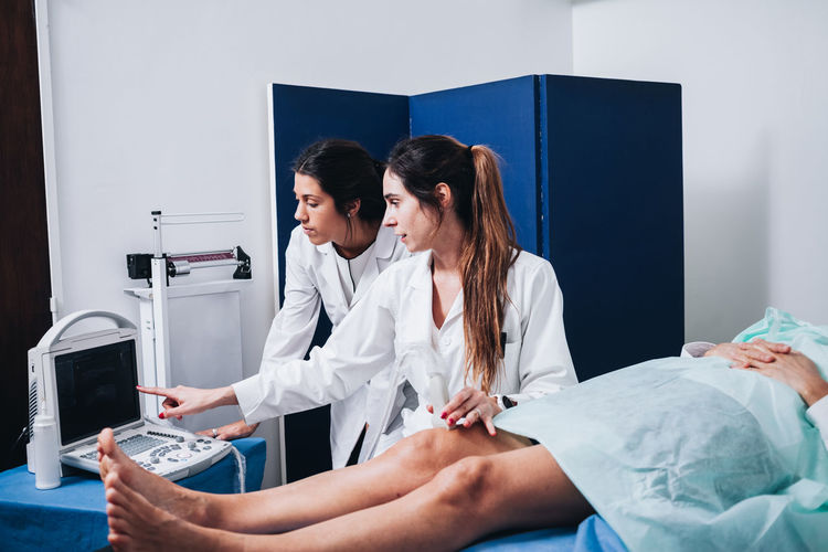Two female doctors are making an ultrasound to the legs of a patient that is lying on a stretcher in an hospital