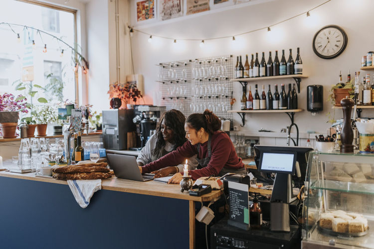 Smiling young female owner discussing over laptop with colleague at checkout in cafe