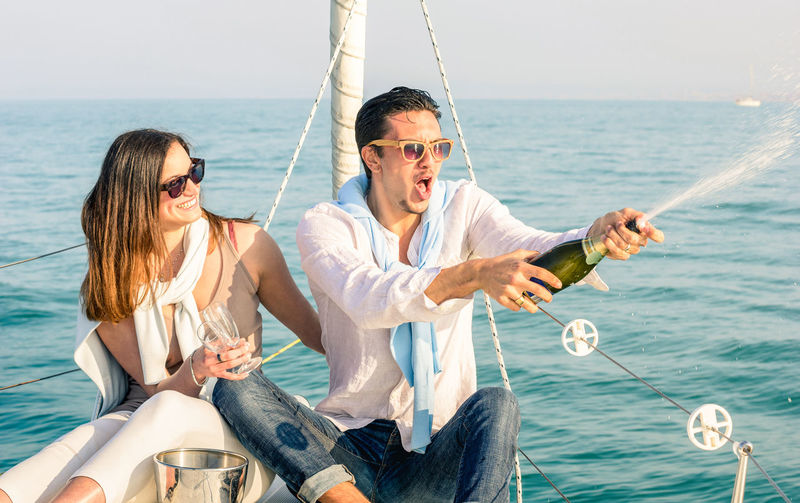 Man spraying champagne with woman holding glasses while sitting on boat in sea
