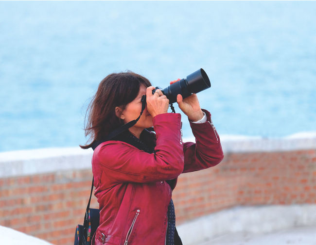 Female photographer in red leather jacket looking thru camera in front of brick a wall