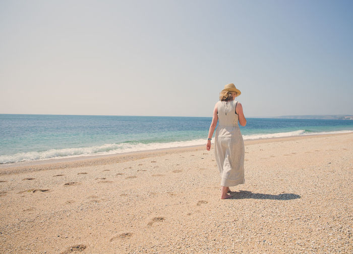 Rear view of mature woman wearing hat while walking at beach against clear sky during sunny day