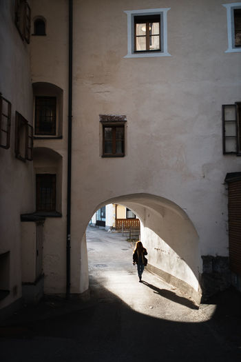 From above back view of unrecognizable female traveler walking through archway of aged stone building in old town