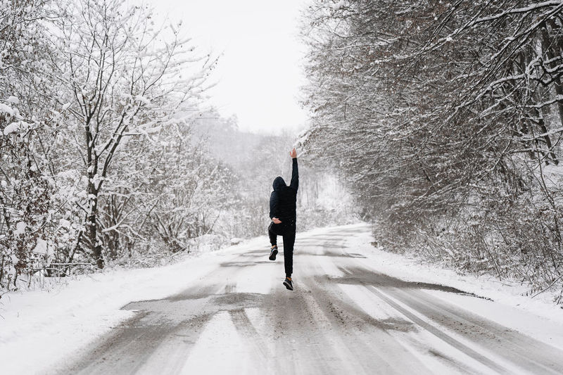 Rear view of man walking and jumping on snow covered road