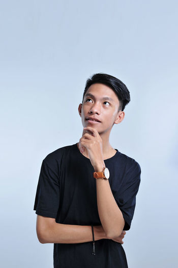 Handsome young asian boy wearing black t-shirt and wristwatch with hand on chin thinking about question, pensive expression. smiling with thoughtful face. doubt concept
