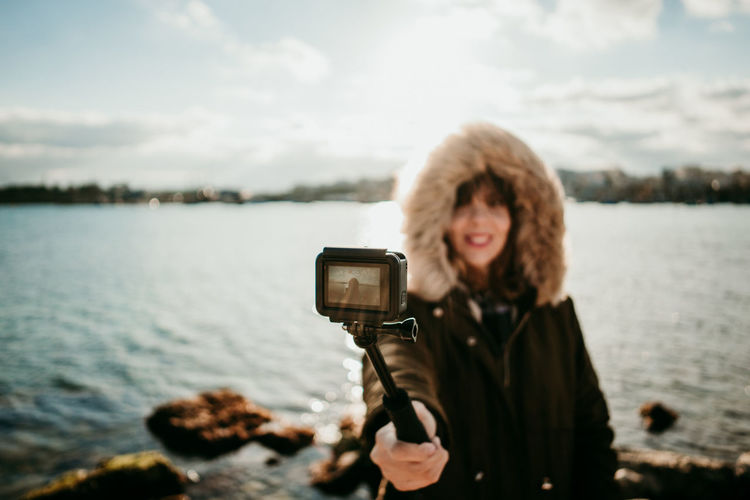 Smiling woman taking selfie with camera while standing against lake