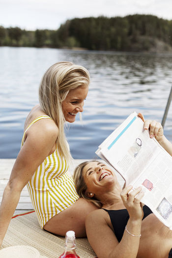 Smiling women in swimsuits reading newspaper at lake