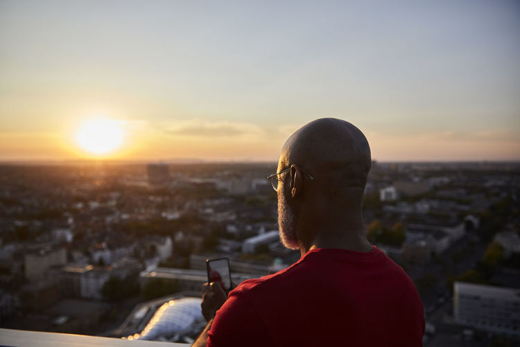 Bald man using smart phone for photographing sunset over city from terrace