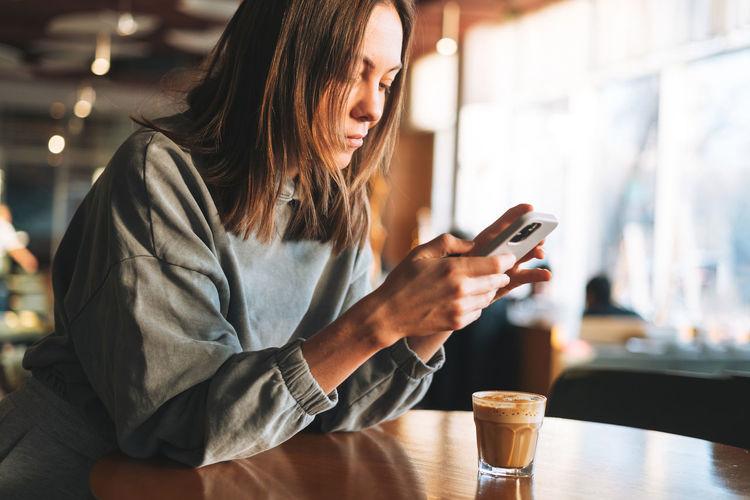 Young woman wearing casual longsleeve with mobile phone in hands with cup of coffee sitting in cafe