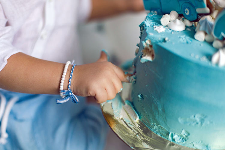 Baby boy in blue pants and shirt standing on the floor next to the cake in blue and white balloons