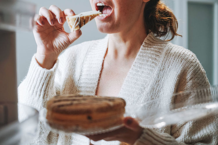 Attractive friendly brunette woman middle age in cozy cardigan eating cake from refrigerator 