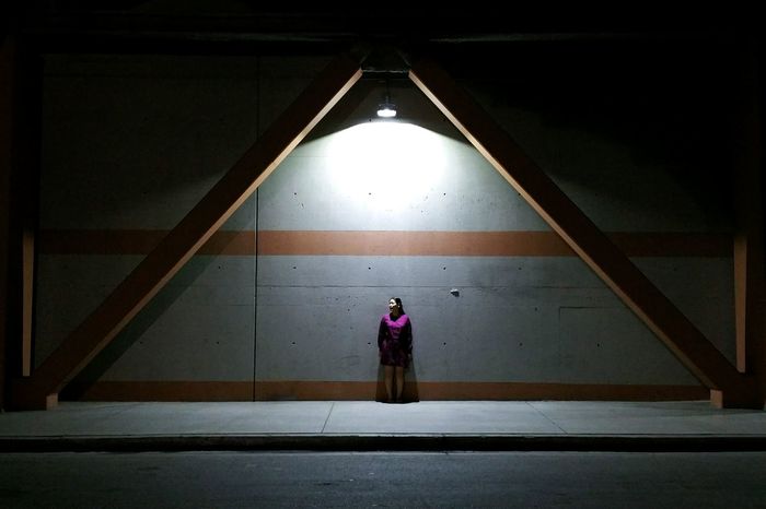Full length of woman standing against wall on sidewalk in city at night