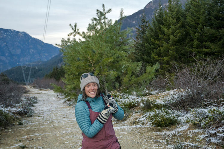 A girl holds a small christmas tree over here shoulder and smiles.