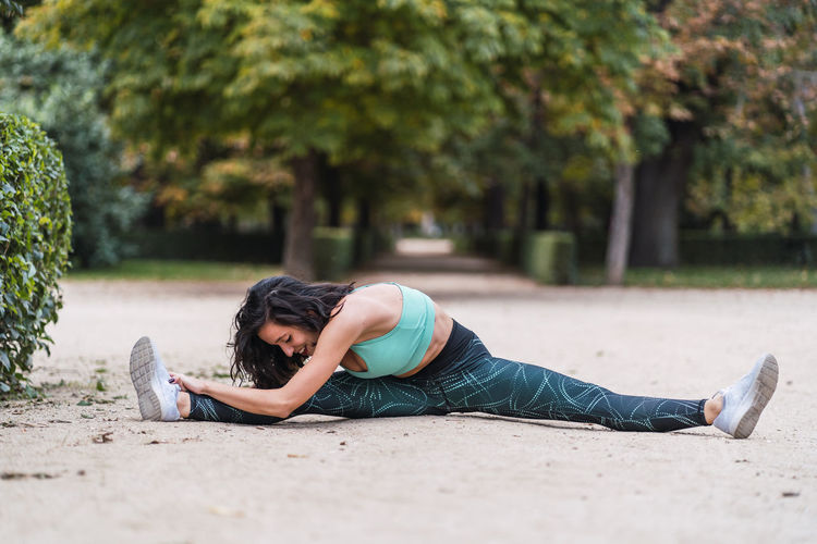 Smiling sportswoman sitting in park and stretching legs while doing splits and warming up during workout